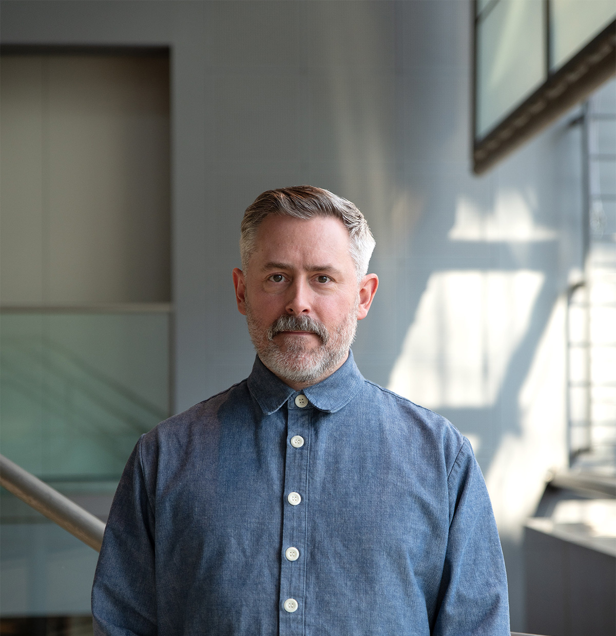 Portrait photo of Martin Hargreaves. A White man with short grey hair and beard wearing a blue button up shirt stadning in a grey room with light coming in from an unseen window