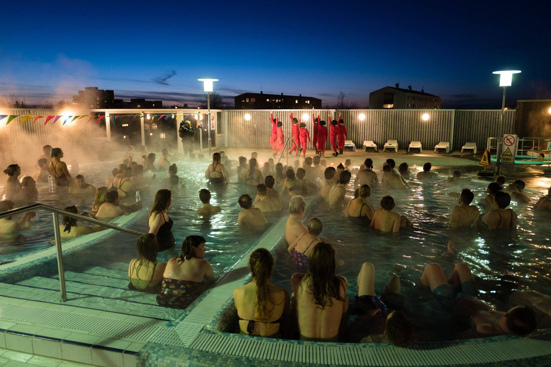 Photo of Reykjavik Dance Festival. Audience seated in hot pools in the dark, with steam rising. Dancers at front of pools in red suits.